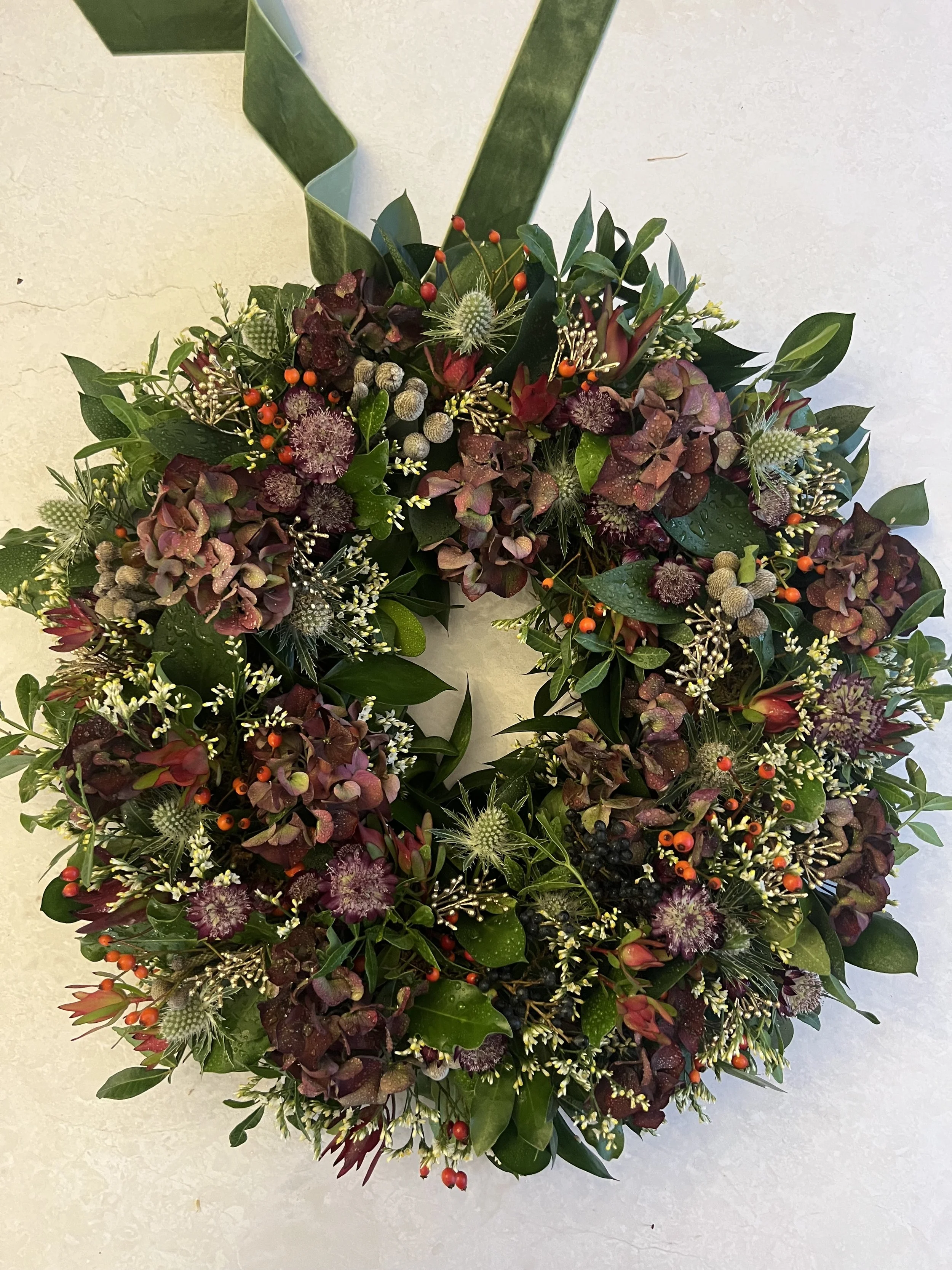 A decorative wreath made of various green leaves and small flowers, with accents of red, purple, white, and orange berries, arranged on a plain light-colored background.