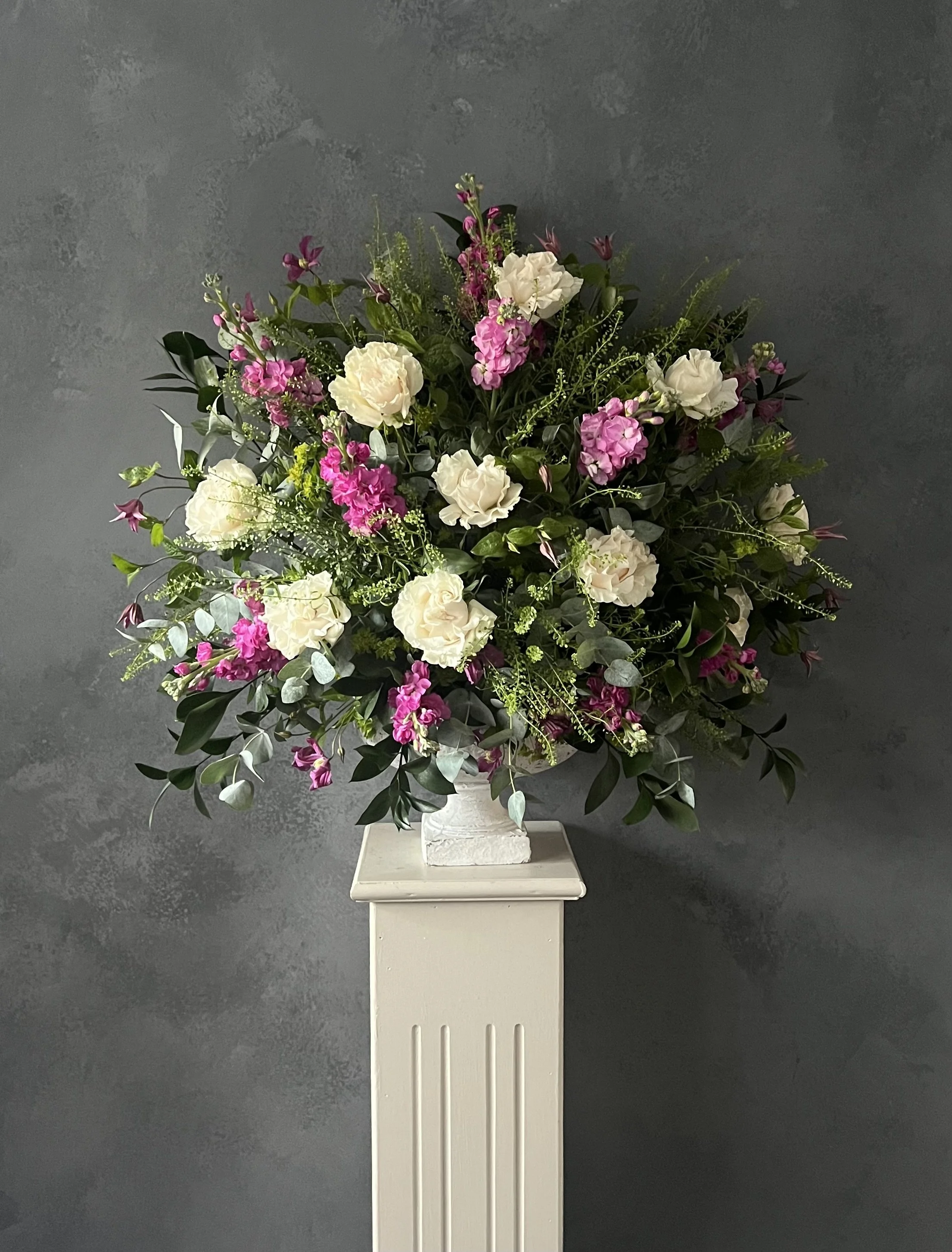 A large floral arrangement with white and purple flowers on a white pedestal against a dark gray textured wall.