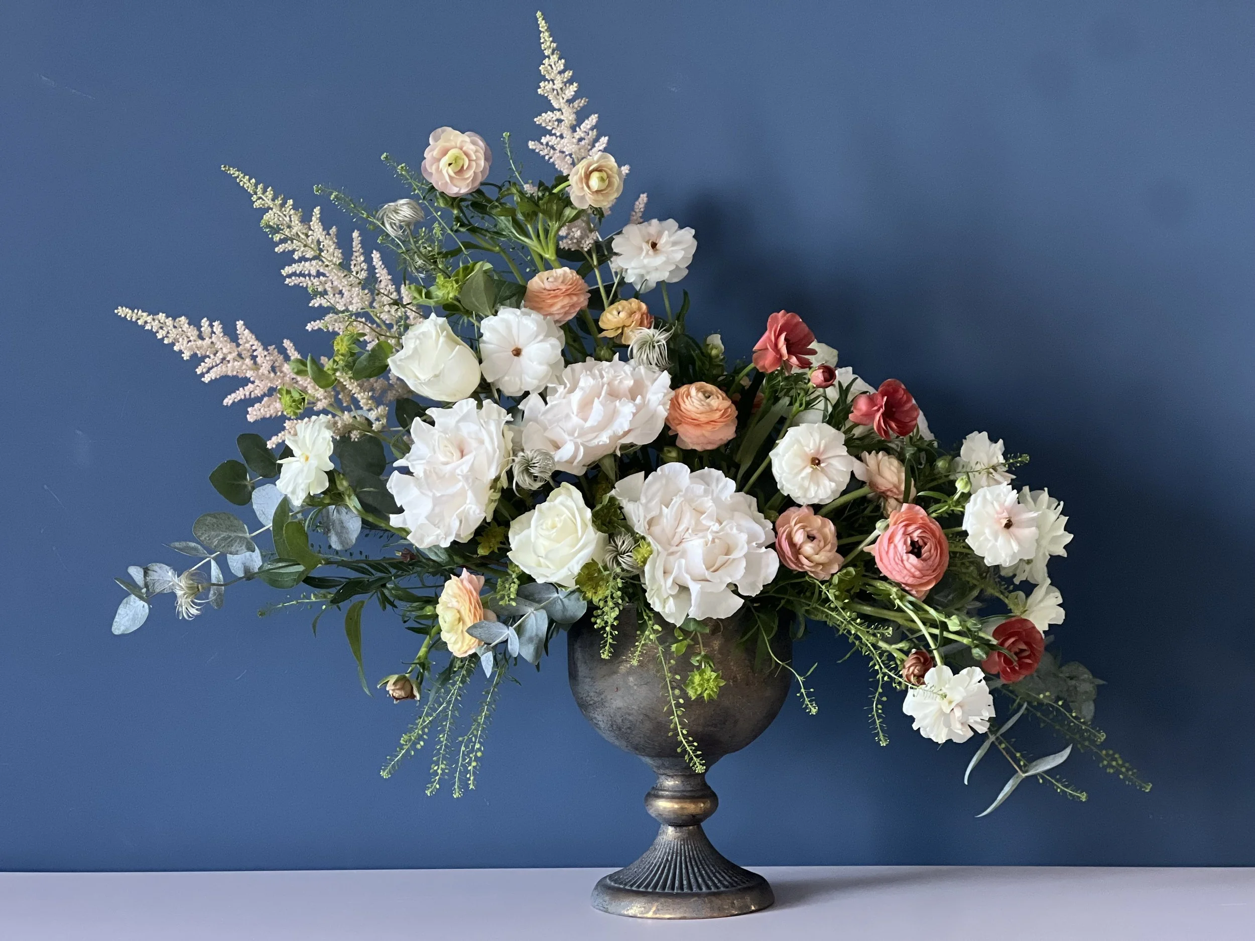 A large floral arrangement in an ornate metal vase with white, pink, and peach roses, white and pink ranunculus, and greenery against a blue background.