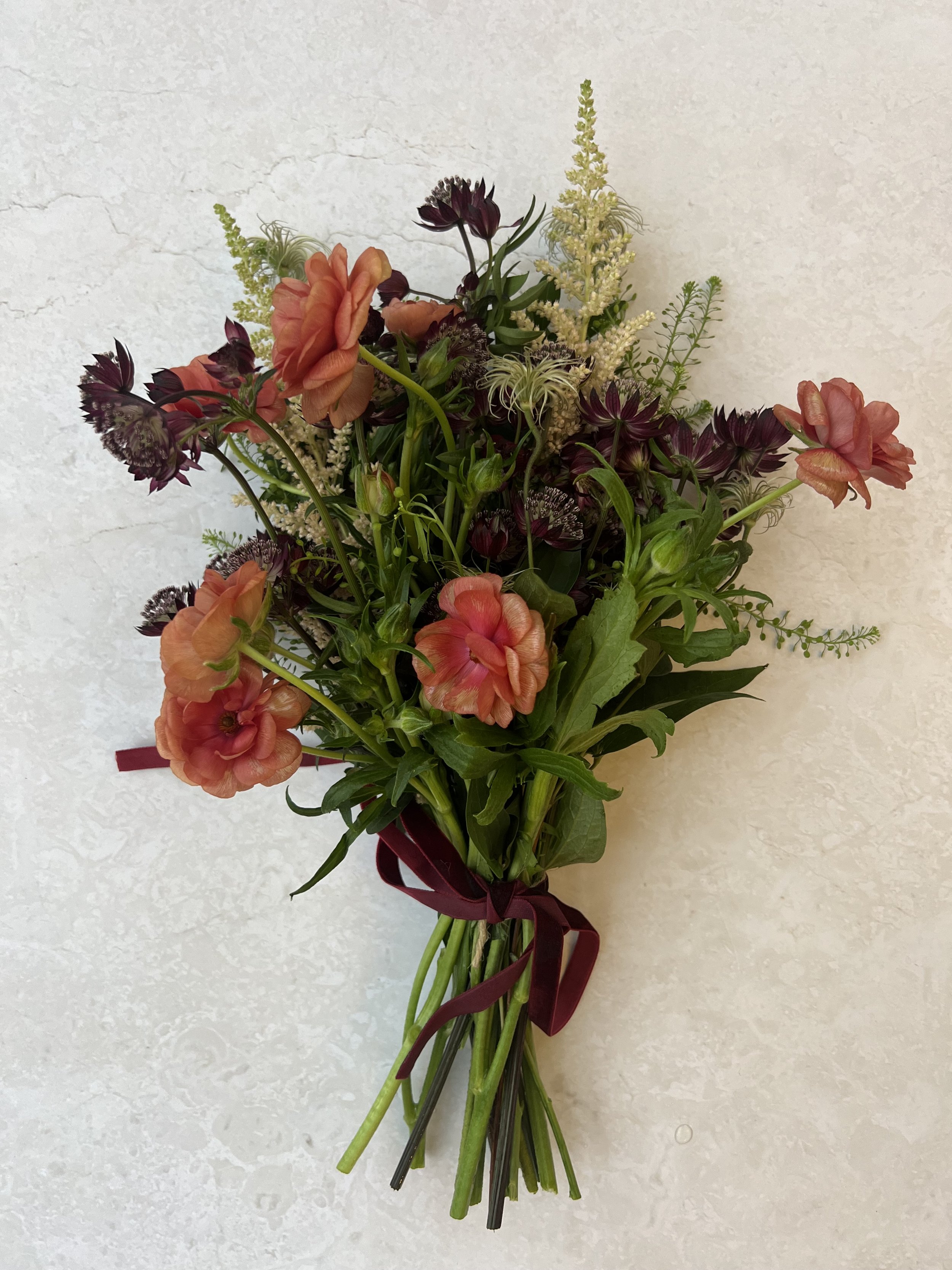 A bouquet of mixed flowers including peach roses, dark purple flowers, cream-colored accents, and green foliage tied with a dark red ribbon.