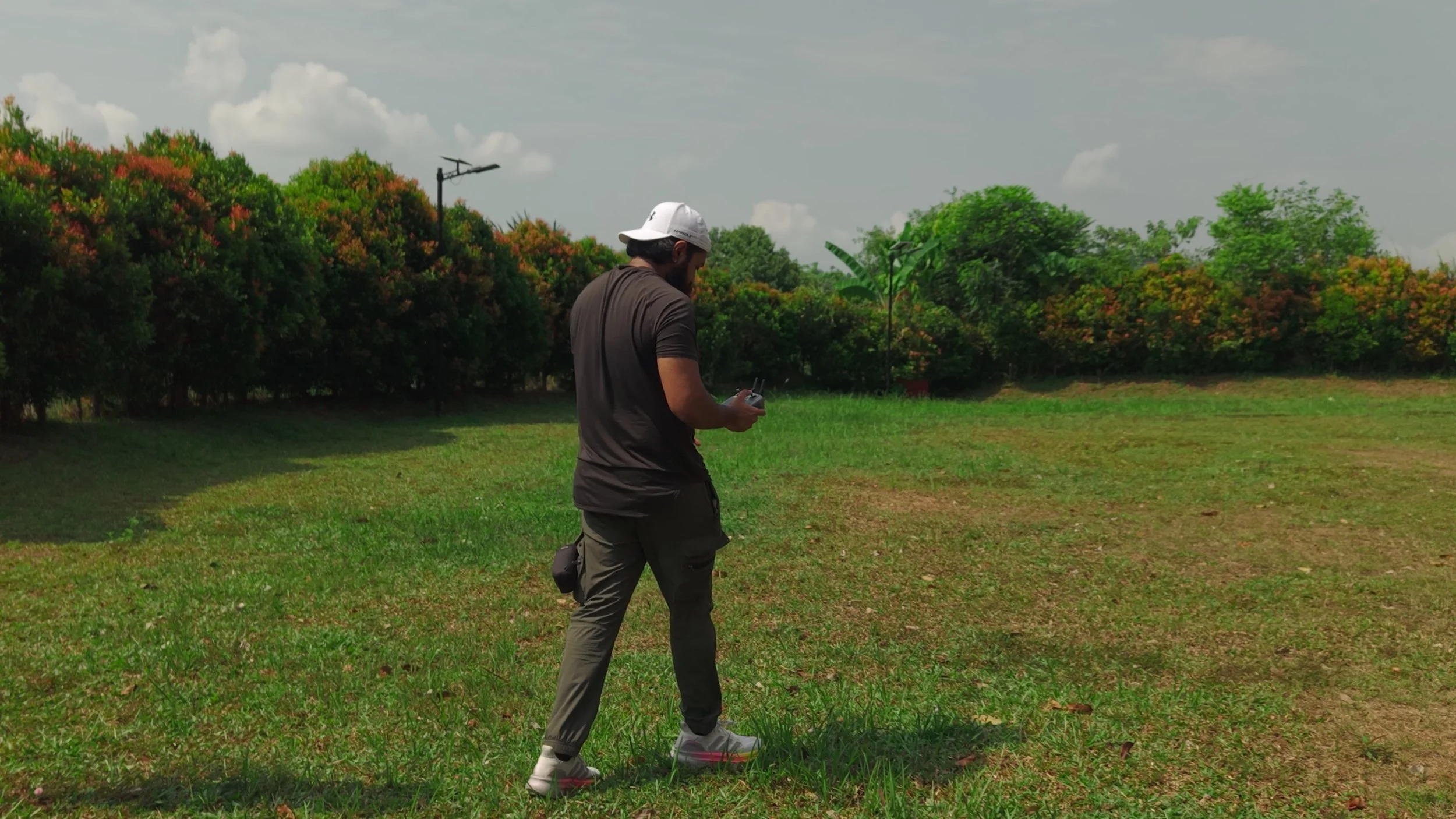 A man walking outdoors in a park-like area, holding a remote control, with trees and bushes in the background, and a partly cloudy sky overhead.