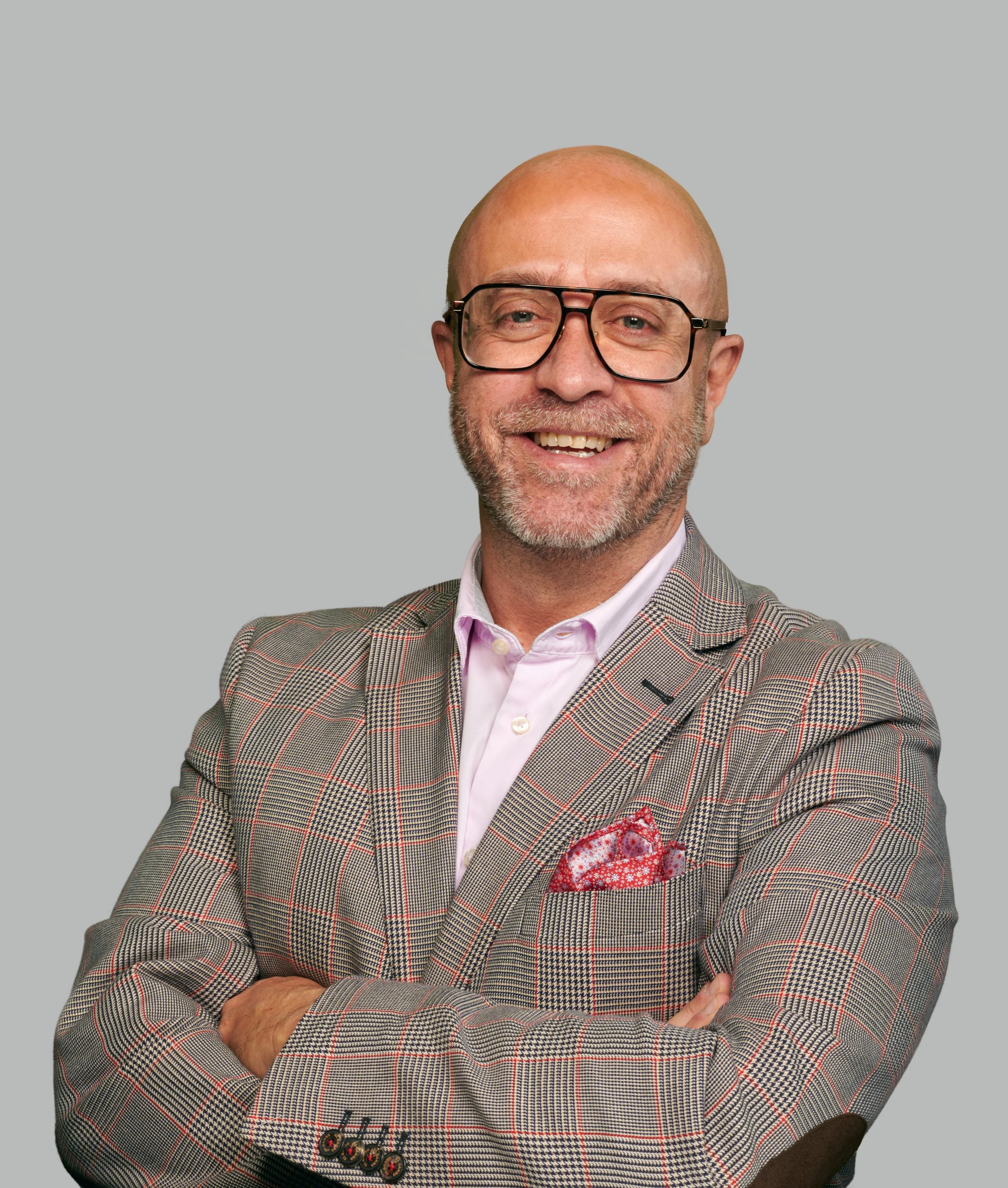 Un homme souriant portant des lunettes, une veste à carreaux, une chemise rose et une poche en mouchoir rouge à pois blancs, avec un fond gris clair.