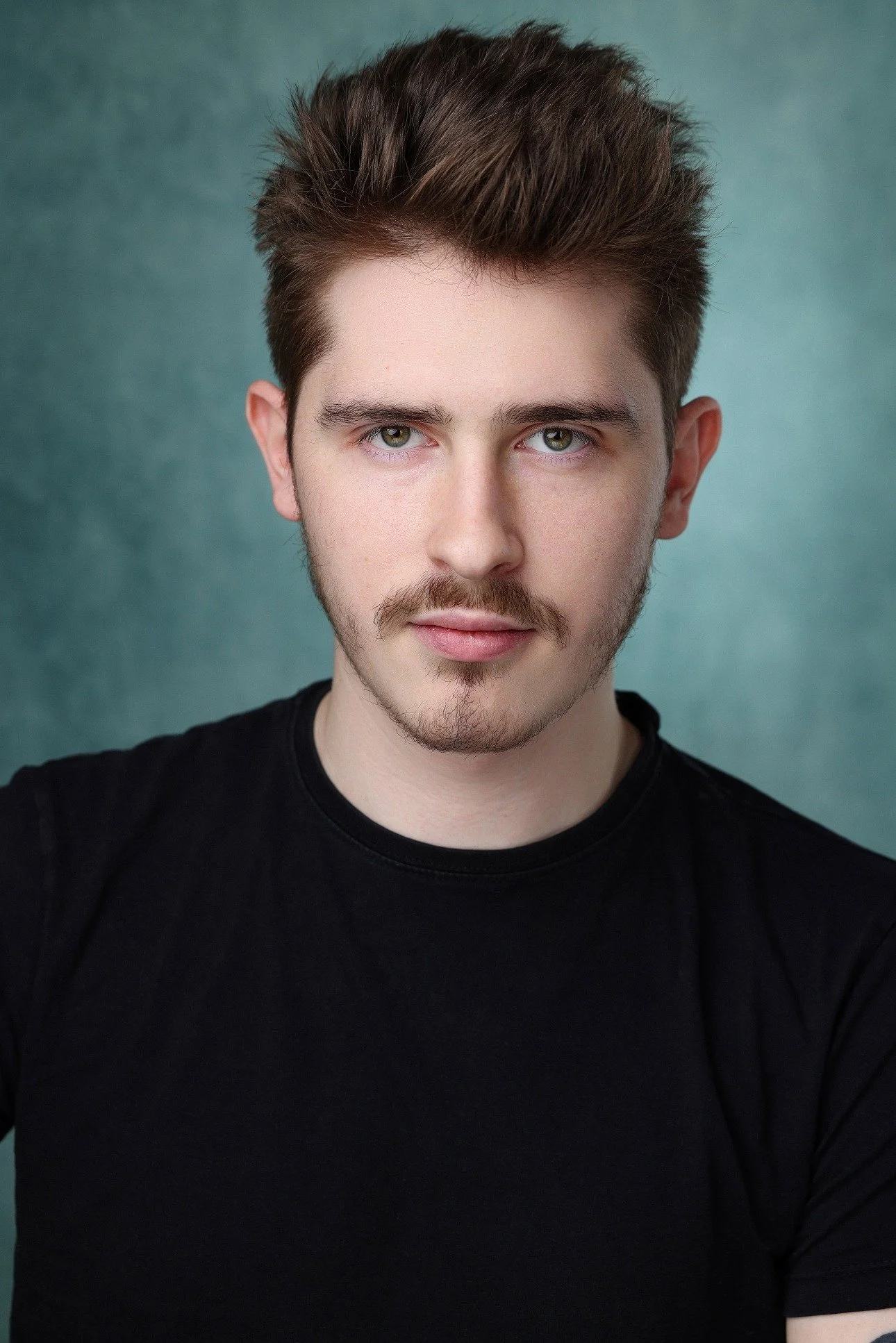 Portrait of a young man with styled brown hair, a mustache, and stubble, wearing a black shirt against a gray background.