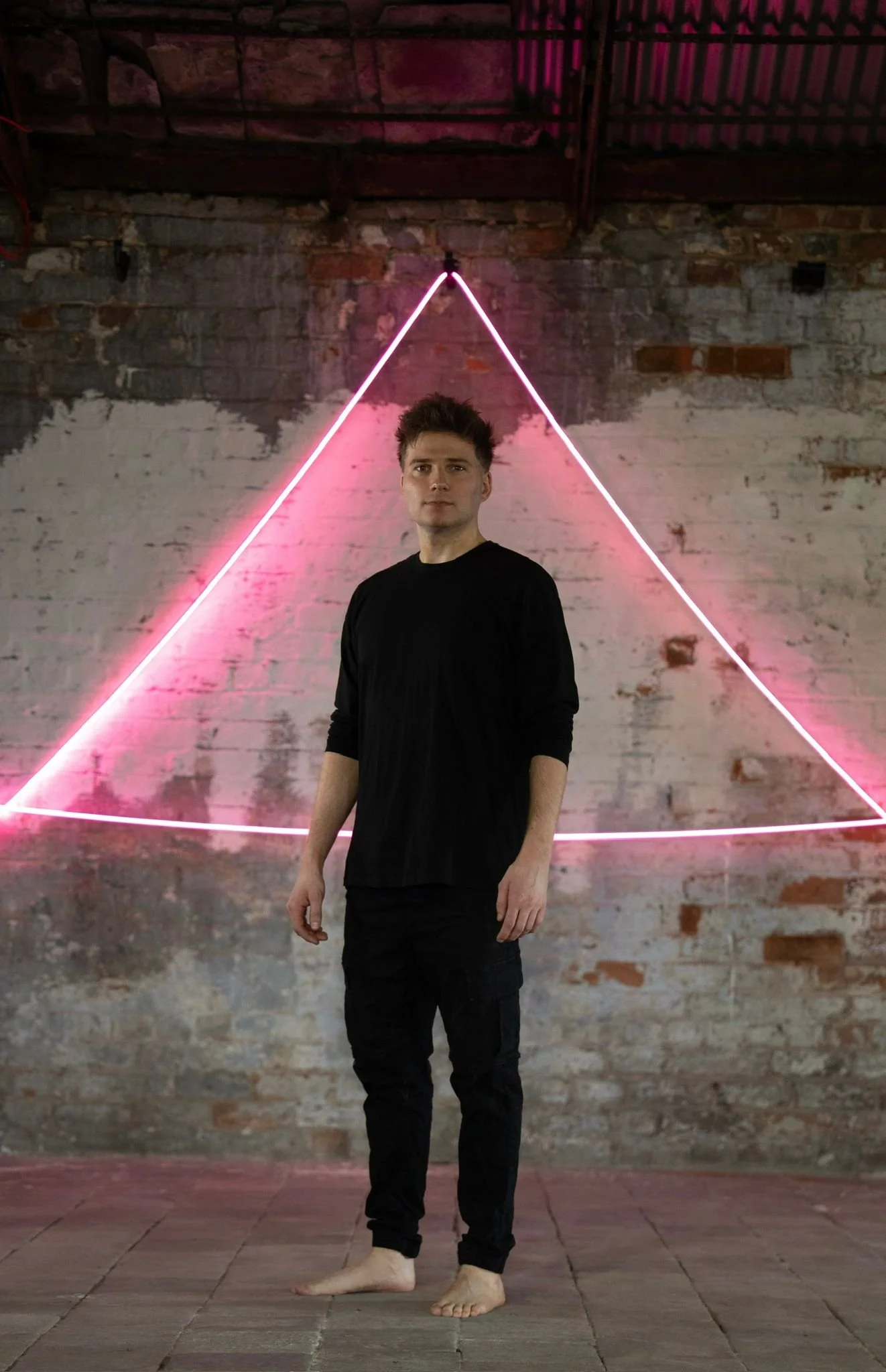 A young man in black clothing standing barefoot on a brick floor in front of a brick wall with pink neon triangle lighting behind him.