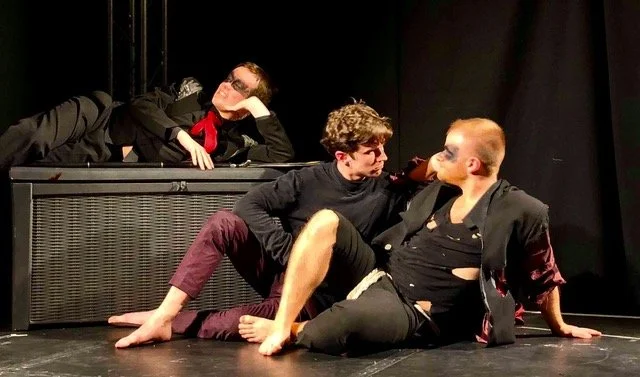 Three young men performing a scene on a stage, two seated on the floor facing each other and one lying on a ledge behind them, all engaged in expressive acting.