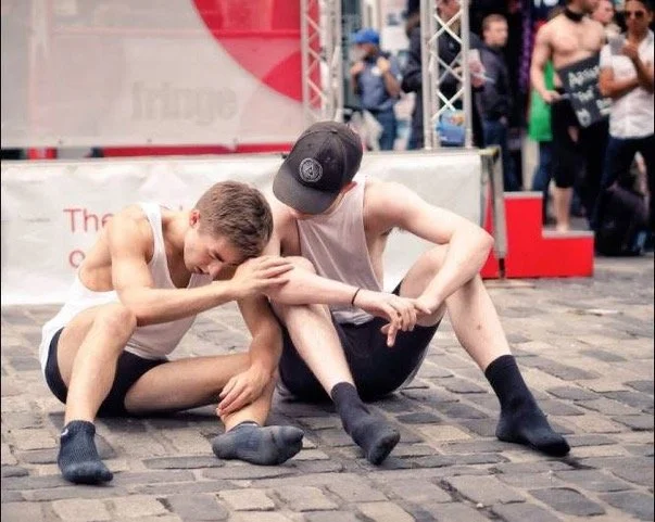 Two young men sitting on a cobblestone street, one comforting the other with his arm around him.