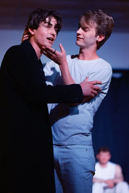 Two men are engaged in a dramatic scene, with one man touching the other's chin and the other looking emotional. A third person is blurred in the background.