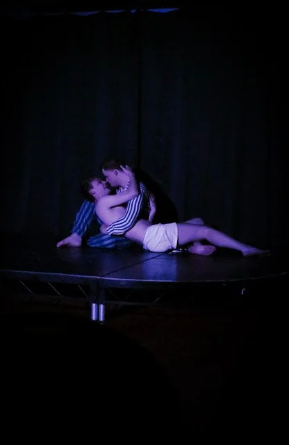 Two young children performing a dance or dramatic scene on a dark stage with a black curtain backdrop.