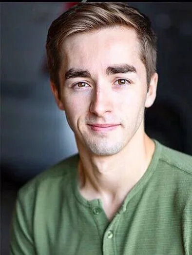 A young man with short brown hair, wearing a green shirt, looking at the camera and smiling slightly.