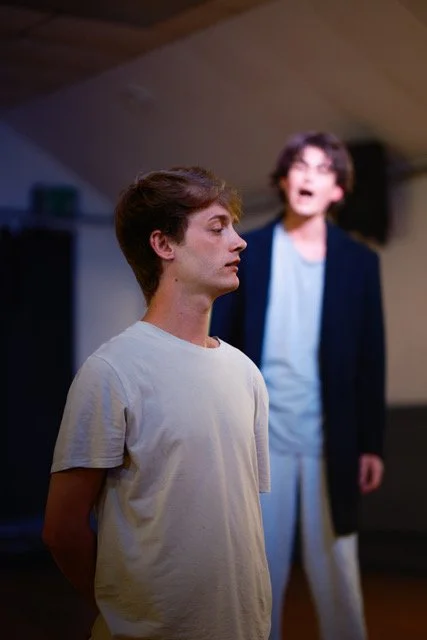 A young man with closed eyes and a neutral expression stands in the foreground, while a woman with glasses, open mouth, and hand gesturing stands in the background, inside a room with a sloped ceiling.