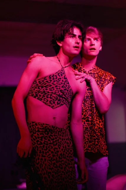 Two mannequins dressed in leopard print clothing, one in a crop top and skirt, the other in a sleeveless top, illuminated by pink and purple lighting.