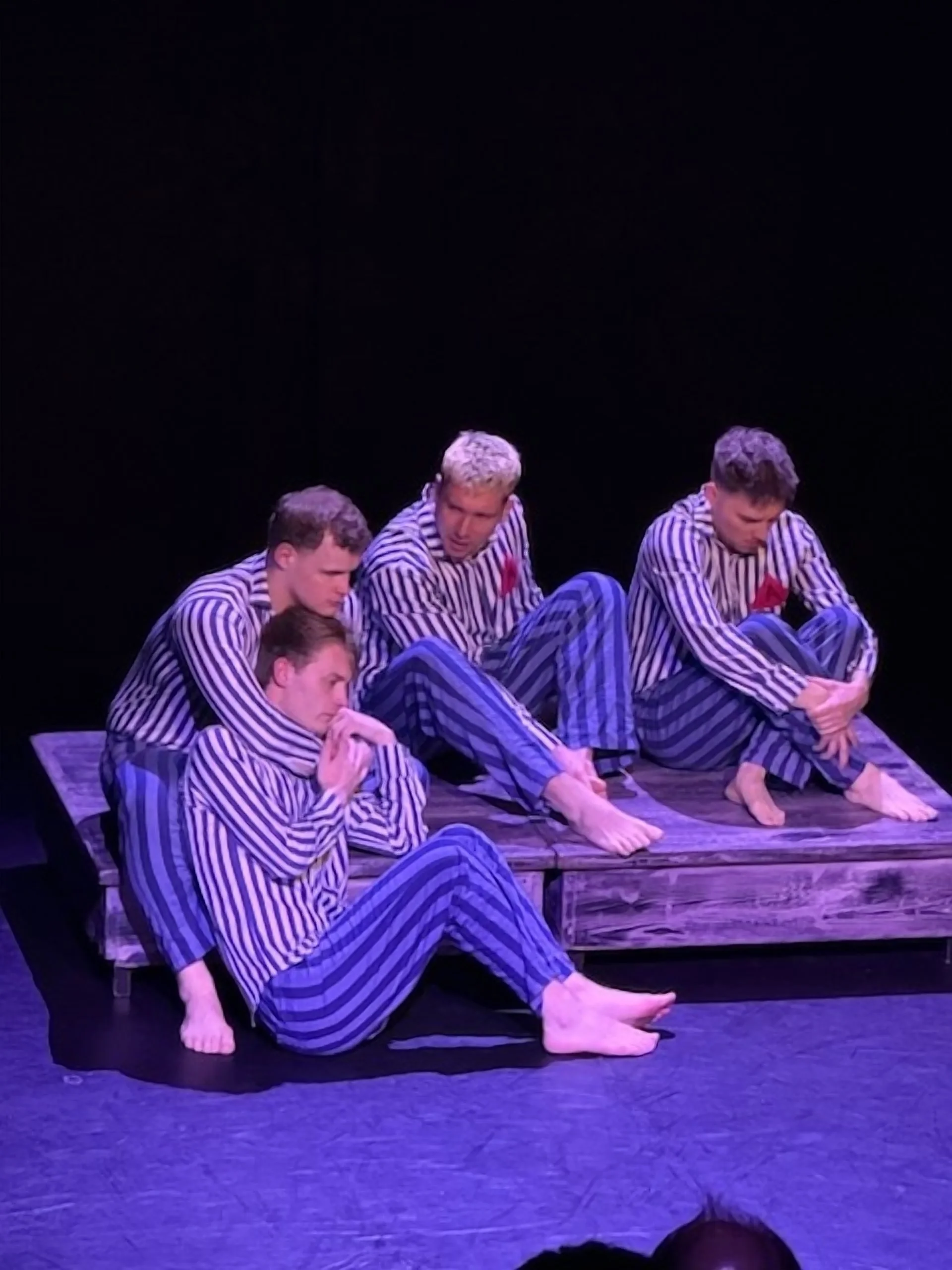 Four young men in striped pajamas sitting and lying on a wooden platform with a black background, performing a theatrical act.