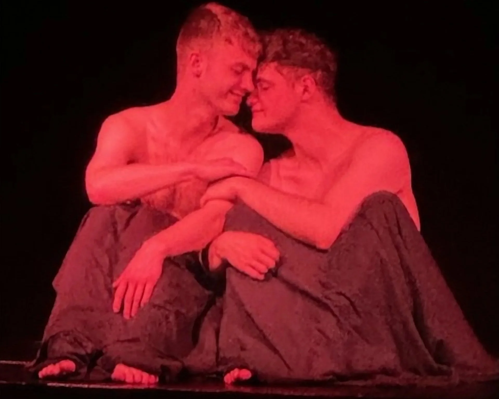 Two shirtless men sitting close together, holding hands, on a stage with a dark background, engaged in an intimate moment.
