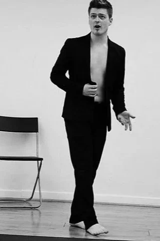 A person with short hair wearing a black blazer and pants, shirtless underneath, standing and gesturing with one hand in a room with a wooden floor and a black chair.