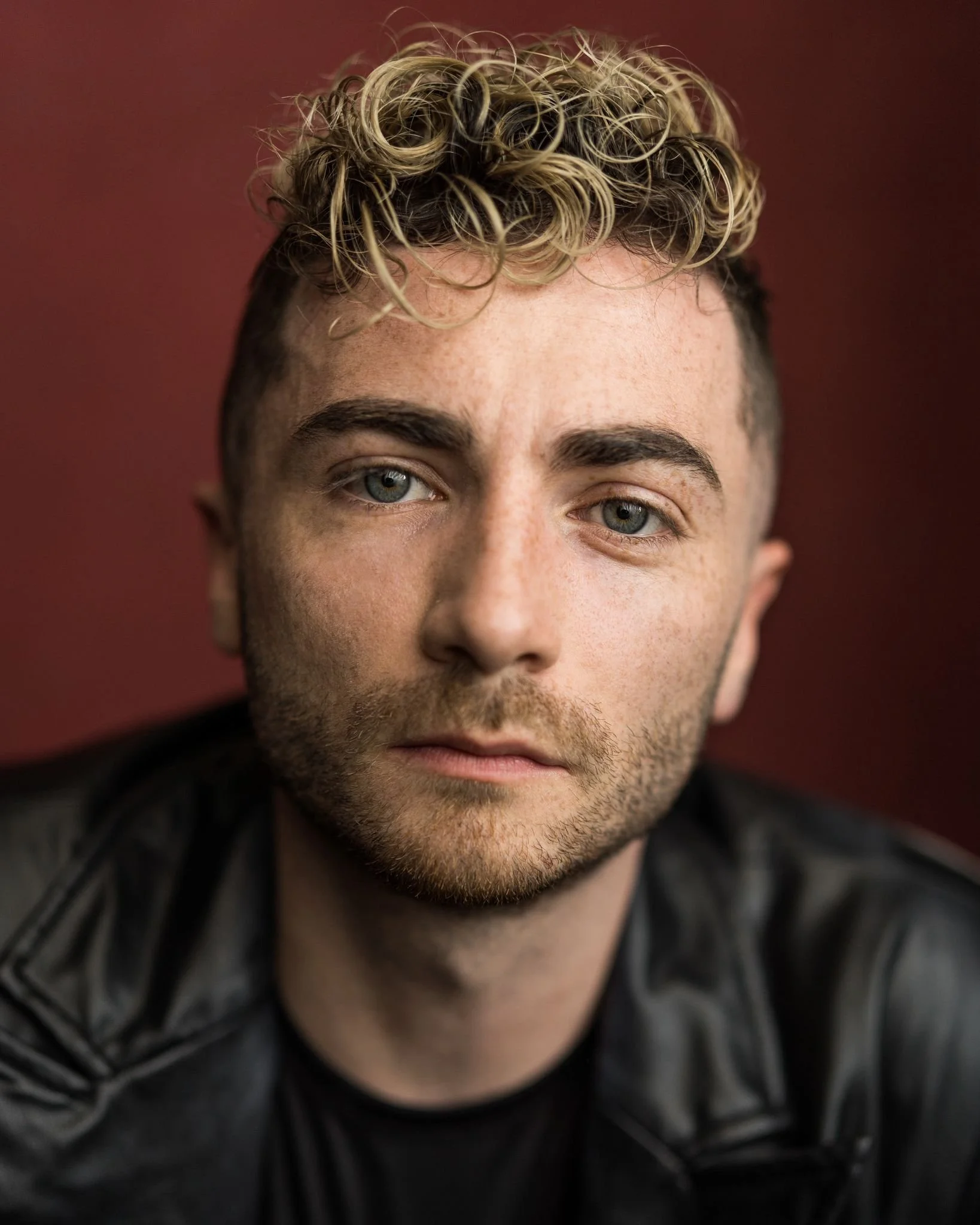 Close-up portrait of a man with curly blonde hair, blue eyes, and facial hair, wearing a black leather jacket against a dark red background.