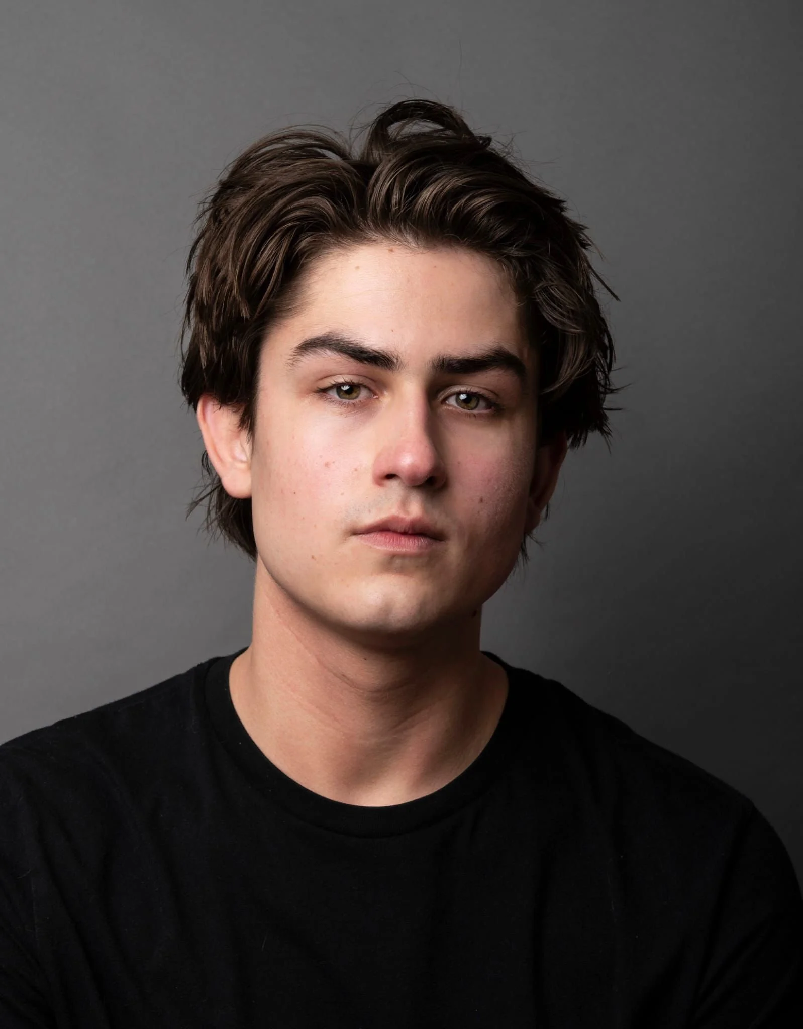 Portrait of a young man with dark brown hair, wearing a black t-shirt, against a gray background.