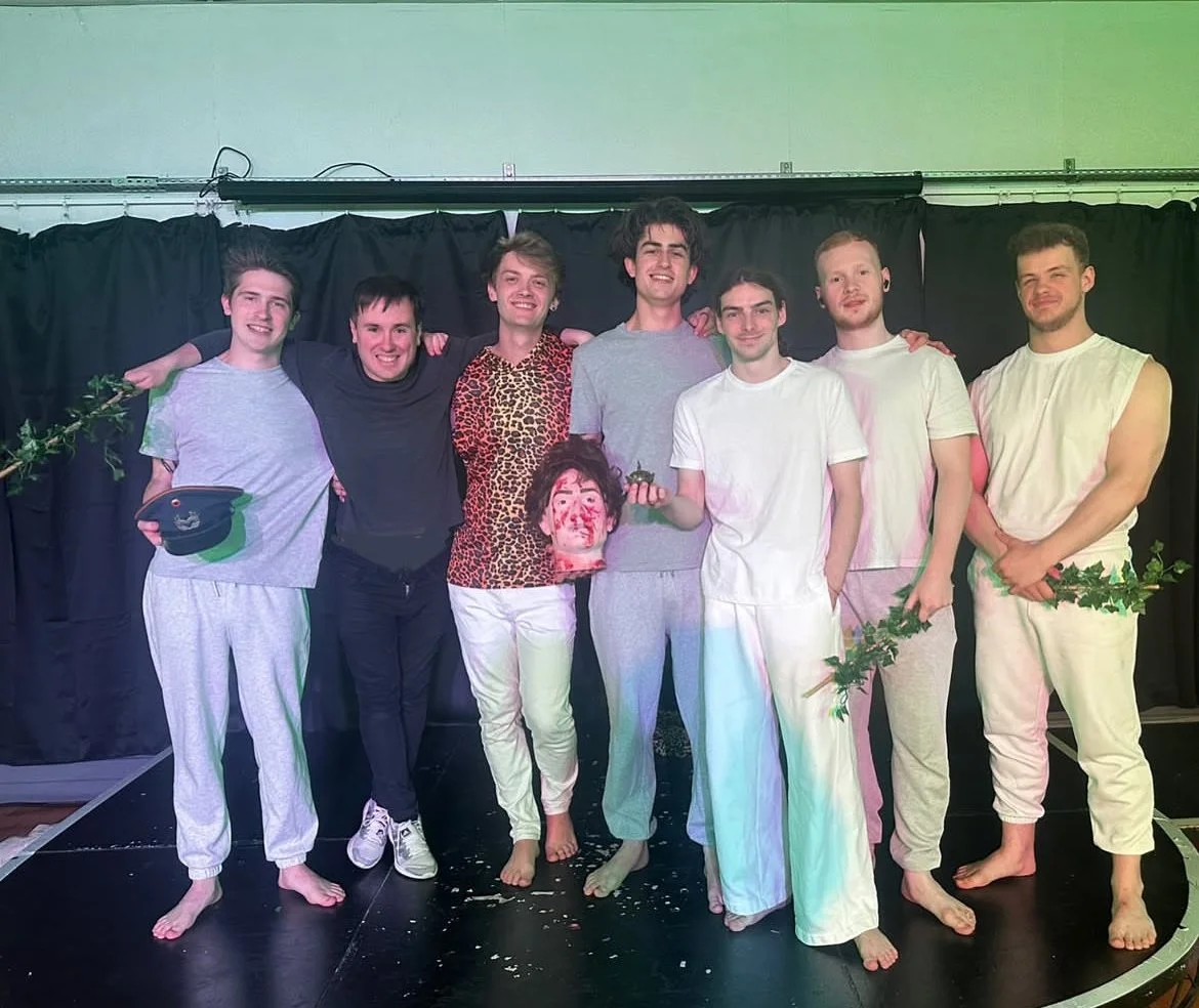 Group of people on stage with black curtain backdrop, some holding props including a military hat, a cast severed head, a small plant. All in casual all white and grey clothing, most barefoot.