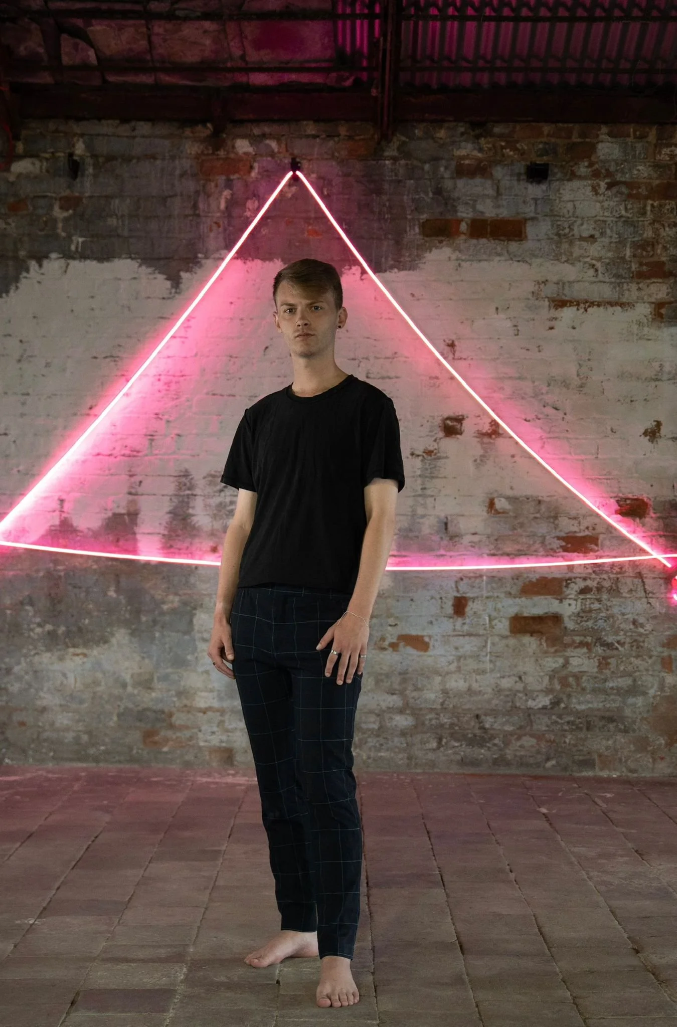 A young person standing barefoot on a tiled floor with a brick wall background illuminated by pink neon lights forming a triangle shape.