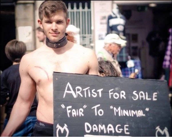 Young man with no shirt, wearing a collar, holding a sign that reads 'Artist for Sale 'Fair' to 'Minimal' Damage' at an outdoor event with people in the background.