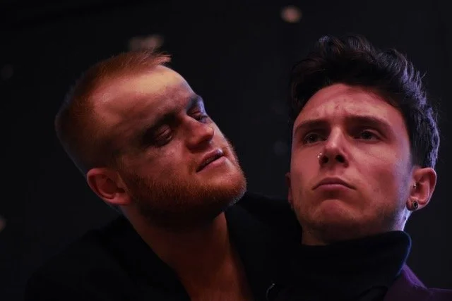 Two young men appear emotional; one has a bruised face, closed eyes, and a pained expression, while the other looks serious and resolute. The scene is dark and intense.