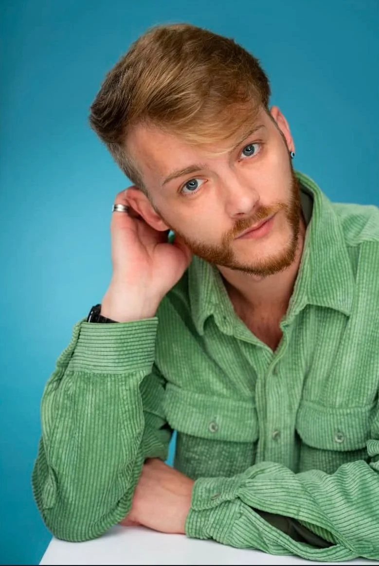 A young man with red hair, blue eyes, and a beard, wearing a green corduroy shirt, resting his head on his hand against a blue background.