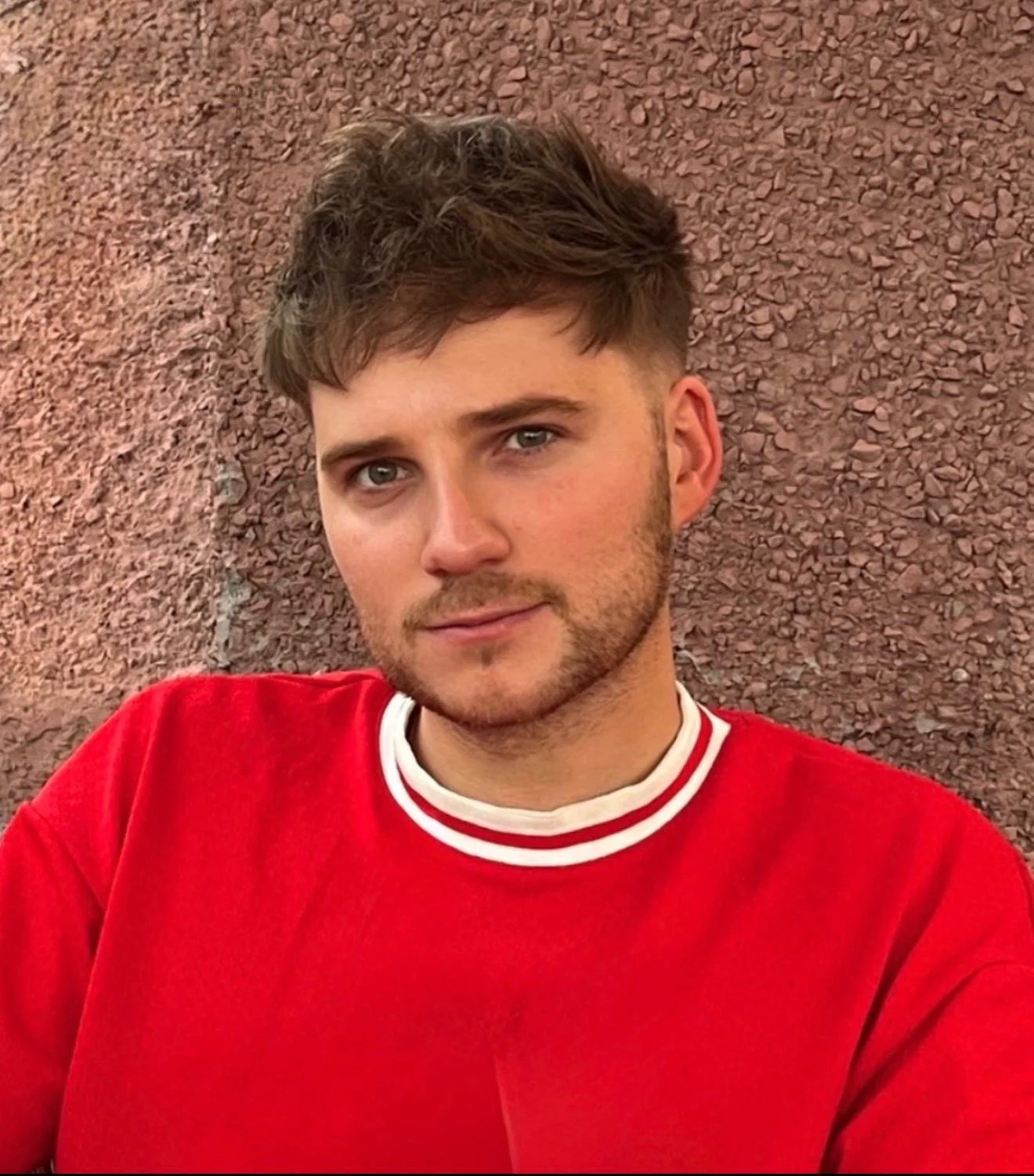 A young man with short brown hair, blue eyes, and facial hair, wearing a red sweatshirt, sitting against a reddish-brown textured wall.