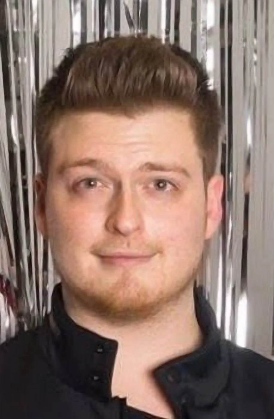 Young man with short brown hair smiling, standing in front of metallic streamers.