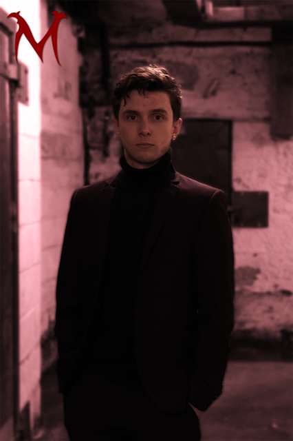 A young man with dark hair and fair skin standing in a dimly lit alley with brick walls, wearing a black turtleneck and blazer, looking directly at the camera.