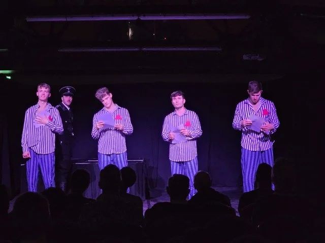Five performers on stage, four dressed in striped pajamas and holding papers, one in a police costume, during a theater or comedy show with an audience watching.