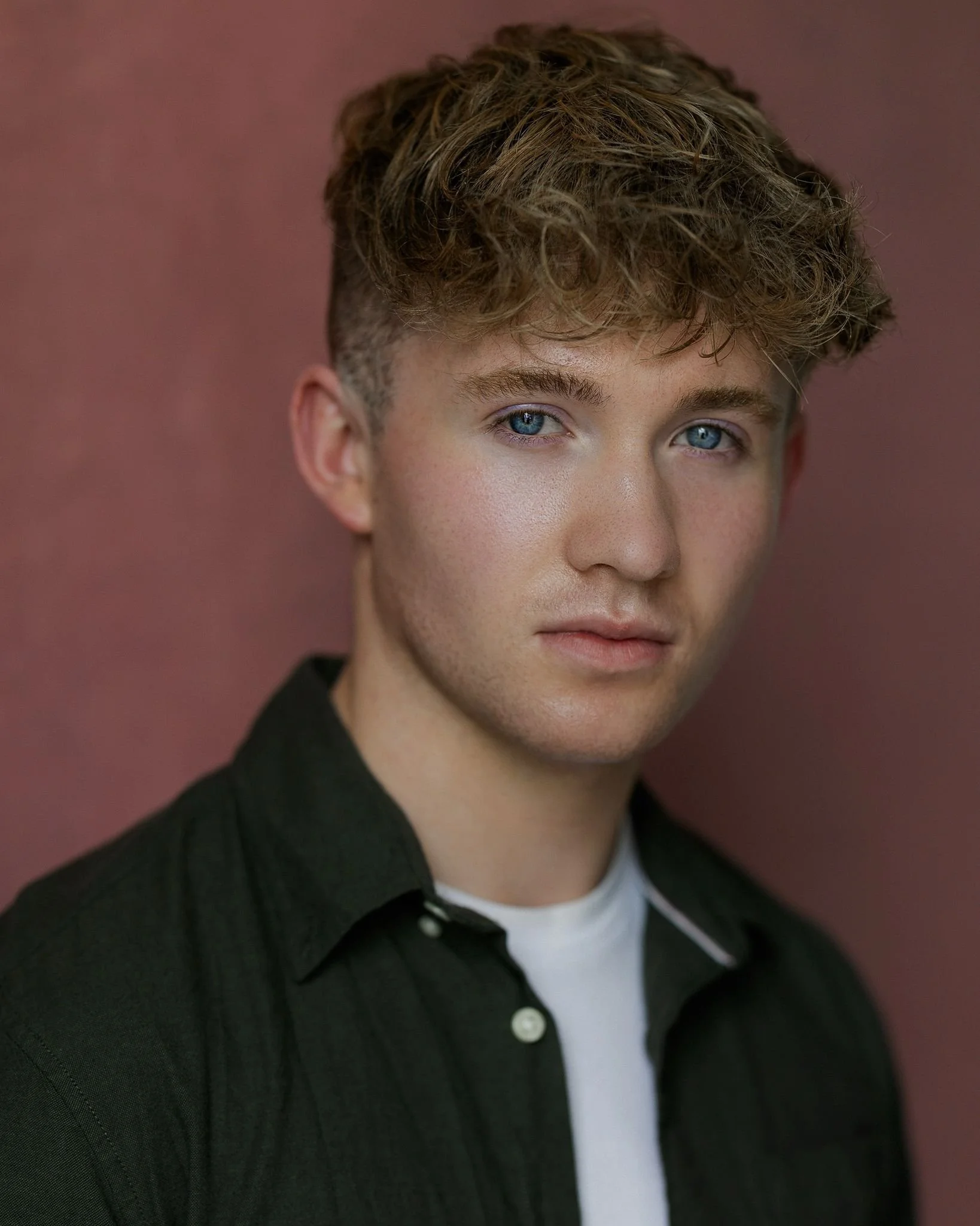 A young man with curly brown hair and blue eyes looking at the camera, wearing a black shirt with a white t-shirt underneath, standing against a reddish-pink background.