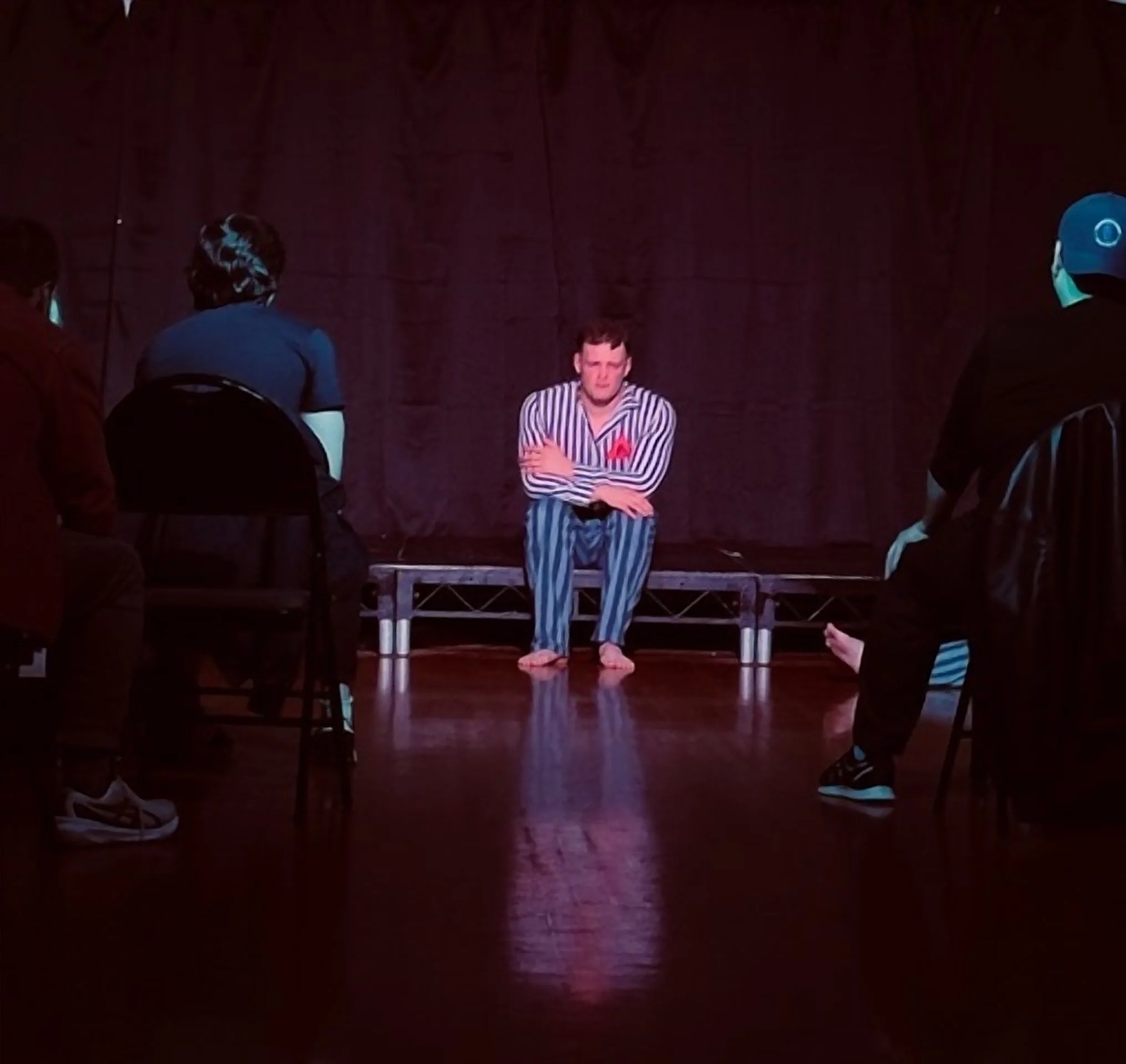 A man in striped pajamas sitting barefoot on a small stage, surrounded by people sitting in chairs, with a dark curtain backdrop.