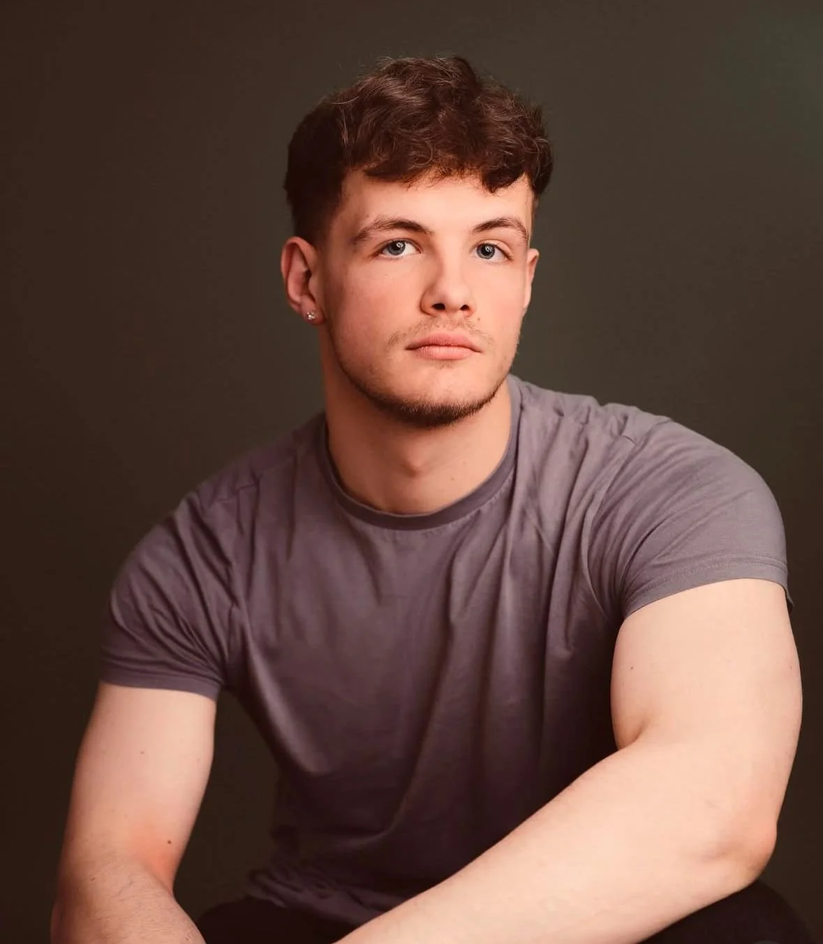A young man with short, wavy brown hair, blue eyes, and a fair complexion, wearing a gray t-shirt, sitting against a dark background.