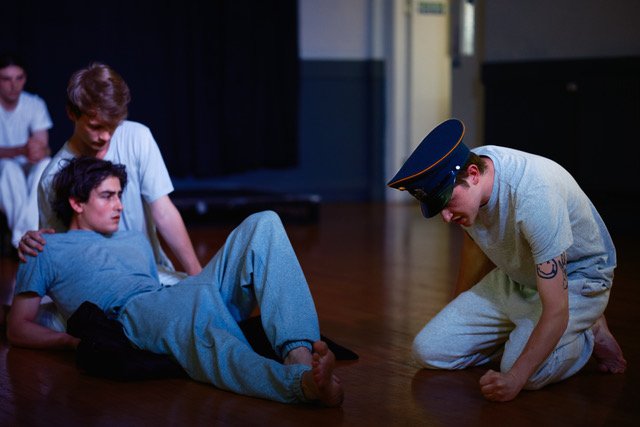Four young men, one lying on the floor, others standing or kneeling around him, in a studio.