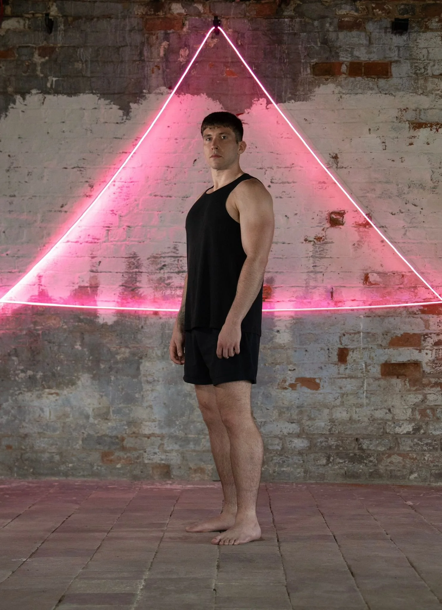 A young man stands barefoot on a tiled floor in front of a brick wall with a pink neon triangle light. He wears a black sleeveless shirt and black shorts, looking at the camera.