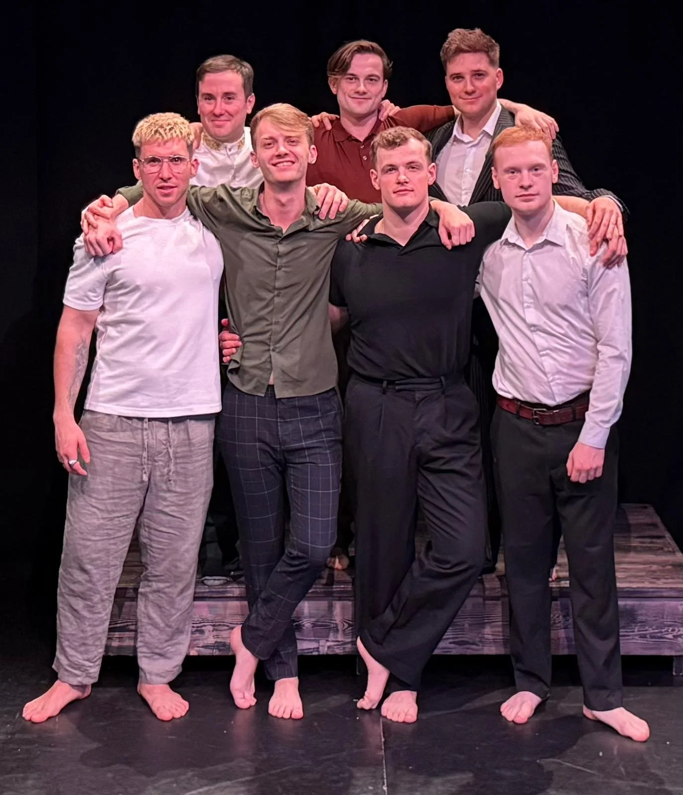 Group of seven young men posing together on a stage with black background, some standing on a wooden platform, dressed in casual and semi-formal clothing, barefoot, smiling and with arms around each other.