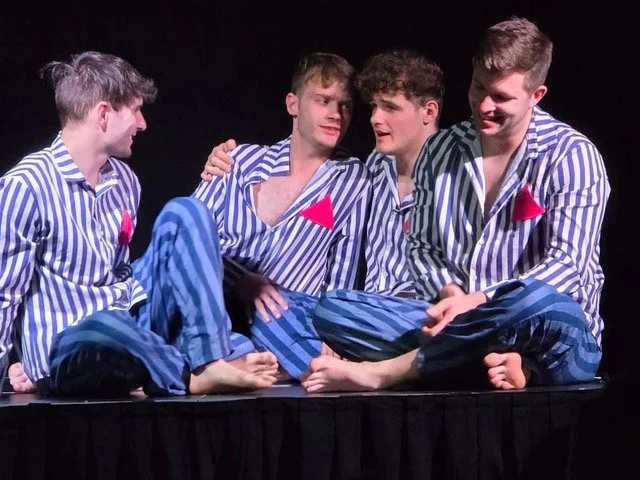 Four young men sitting cross-legged on a stage, wearing matching striped pajamas with red badges, engaged in conversation.