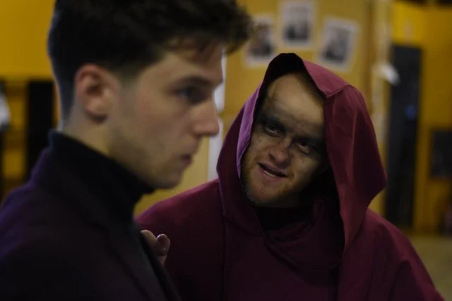 Two men in a tense confrontation, one with a serious expression and the other with face makeup or scars, dressed in a hoodie, in a dimly lit indoor setting with yellow walls and photographs.