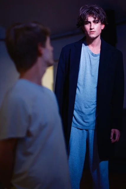 A young man with tousled dark hair wearing a light blue t-shirt and pajama pants, standing in a dimly lit room facing another person with blurred features.