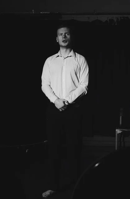 A man stands barefoot on a stage with a black curtain backdrop, wearing a white dress shirt and dark pants, appearing to perform or speak.