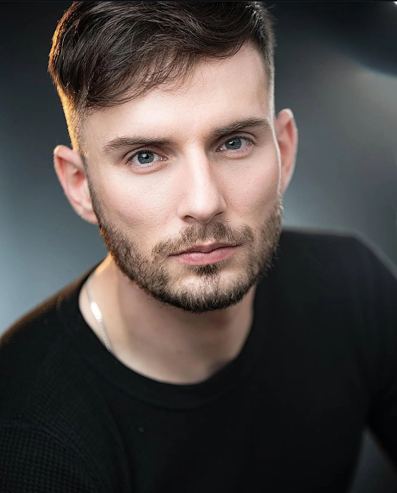 A close-up portrait of a man with short brown hair, blue eyes, and a beard, wearing a black shirt and a silver chain necklace against a blurred dark background.