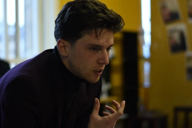 A young man with dark hair and fair skin looking focused with his hand raised, in a room with yellow walls and windows.