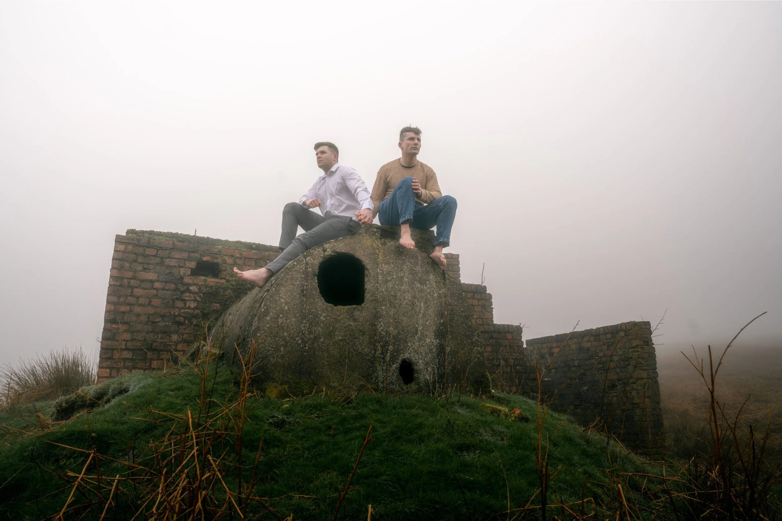 Two men sit on a large, old, stone bunker at a foggy outdoor site with brick ruins in the background.