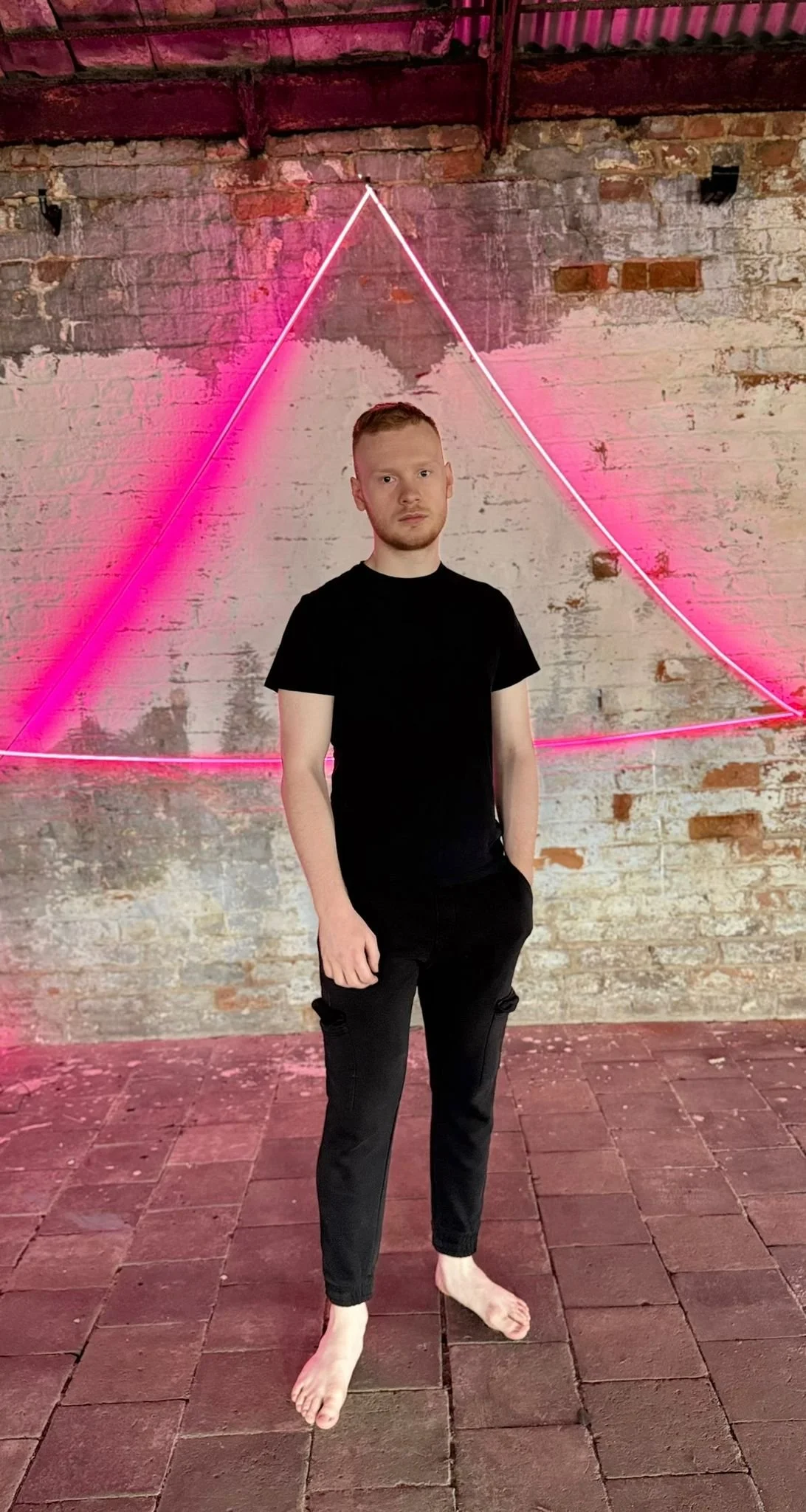 A young man with red hair and beard, wearing a black t-shirt and black pants, standing barefoot in front of a rustic brick wall with pink neon triangle art.