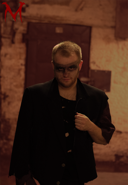 A young man with a moody expression, short blond hair, dark makeup around his eyes, wearing a black jacket over a torn black shirt, standing in a dimly lit, rustic room with exposed brick walls and wooden beams.