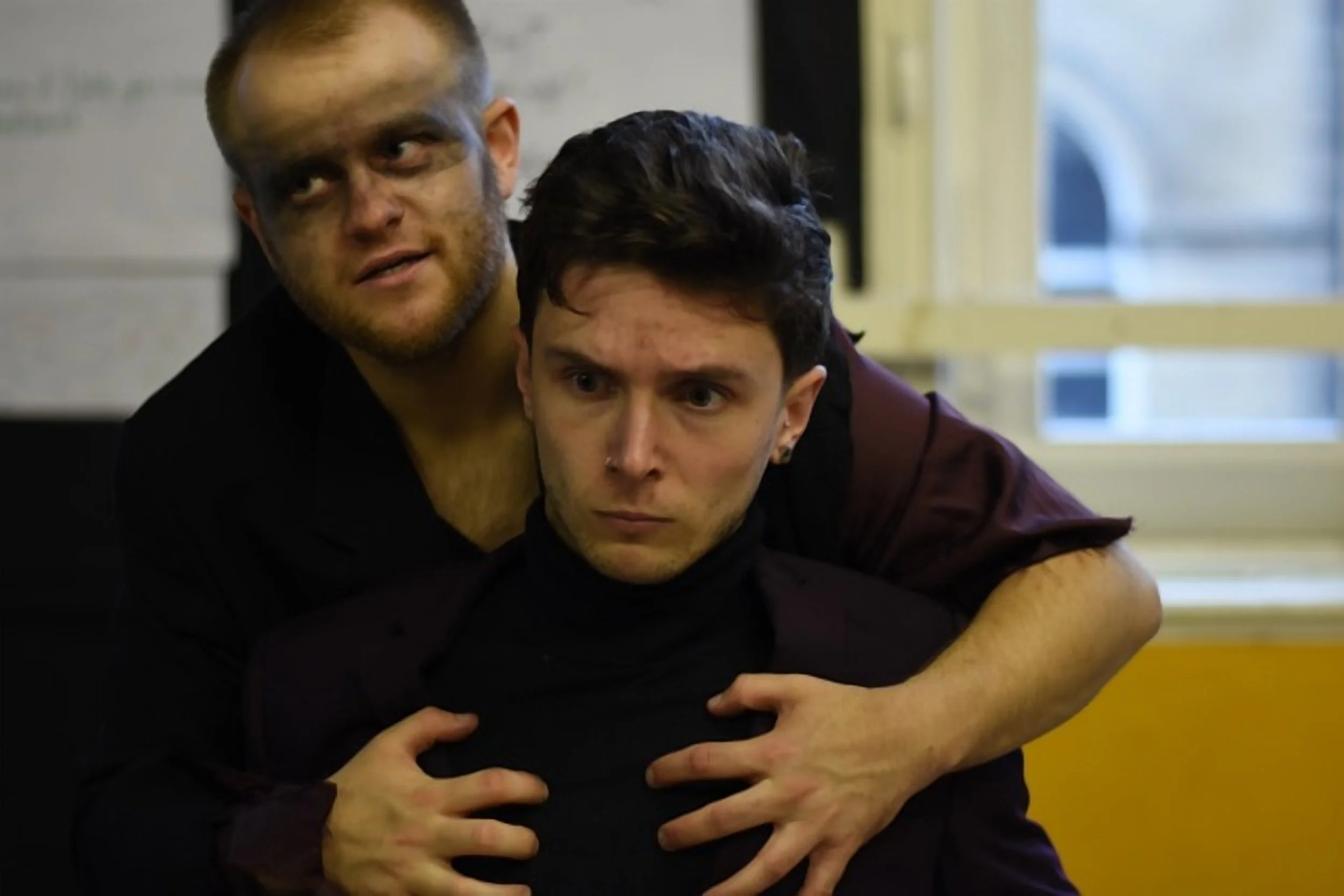 Two men appear to be in a tense situation indoors, with one man grabbing the other from behind. The man in front has short dark hair and a serious expression, while the man behind has light hair and facial hair, and is looking to the side with a concerned expression.
