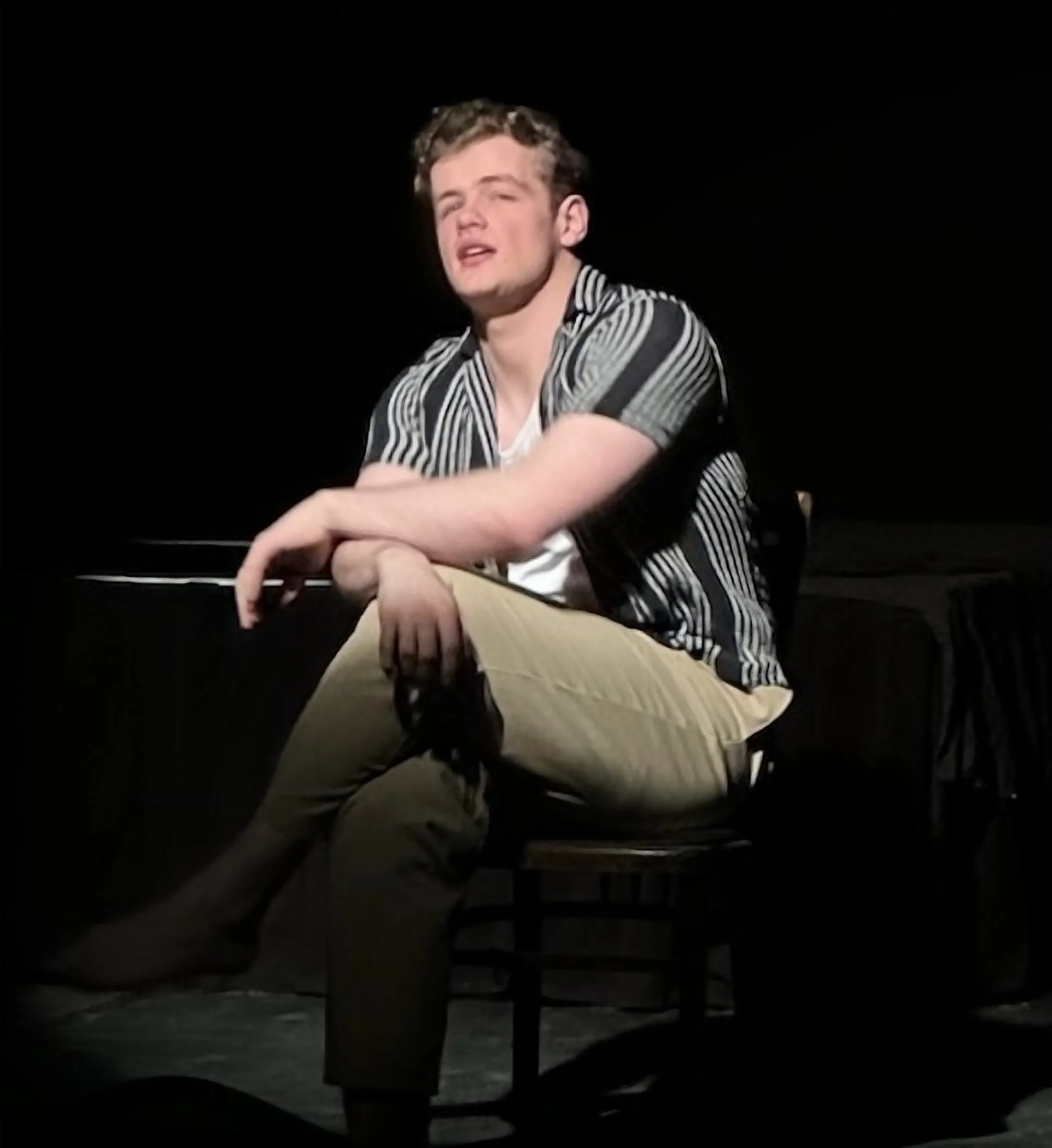 A young man with short curly hair sitting on a wooden chair on a dark stage, wearing a striped short sleeve shirt and beige pants, with a serious or contemplative expression.