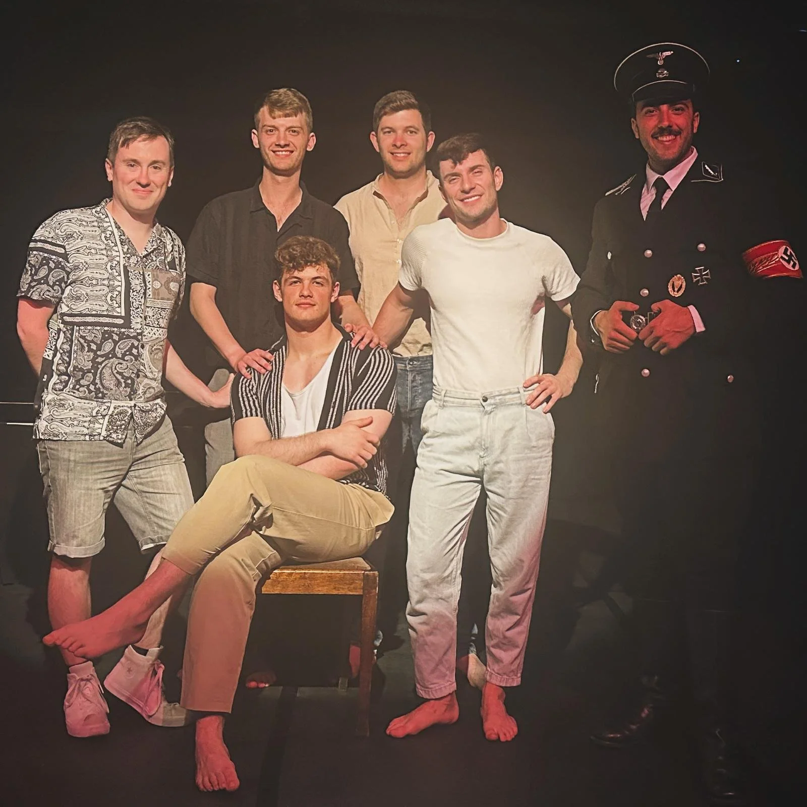 A group of seven young men posing together against a black background. One is dressed in a Nazi uniform, including a hat, armband, and medals. Three are standing, three are seated or kneeling, and one is sitting on a stool barefoot with crossed arms.