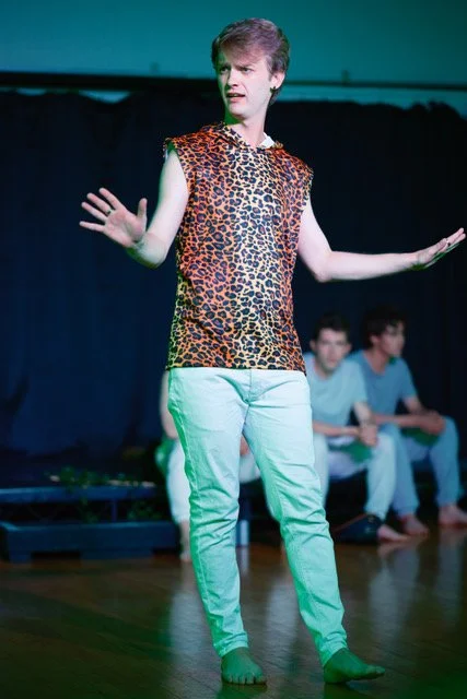 A man with a costume that has a leopard print top, white pants, and no shoes, performing on stage with his hands raised, in front of a curtain with two seated people in the background.