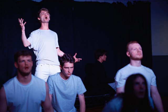 A group of five young people on stage, with one person standing and singing passionately while the others are sitting or standing silent, possibly during a theatrical performance or rehearsal.