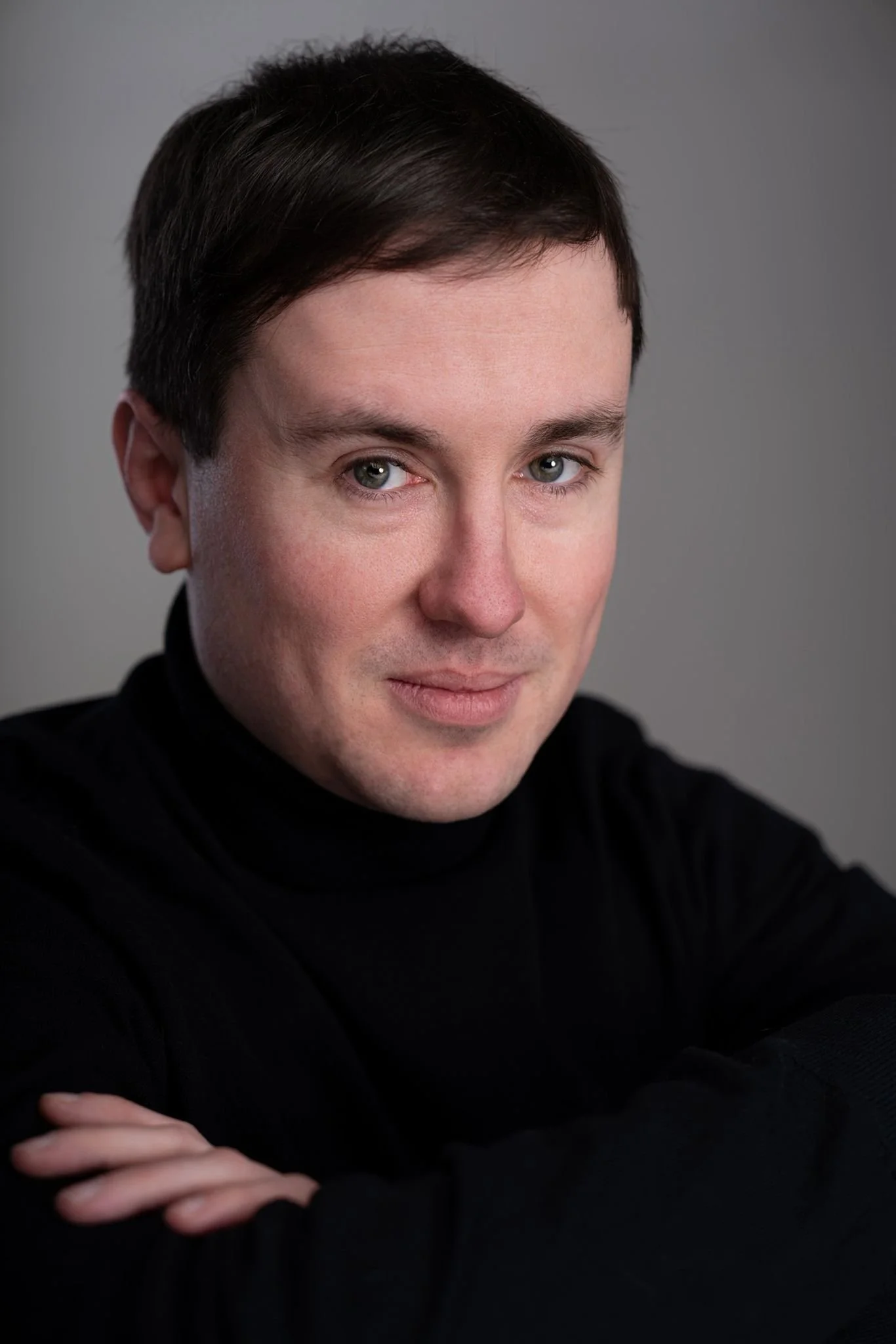 A portrait of a man with short, dark hair wearing a black turtleneck against a gray background.